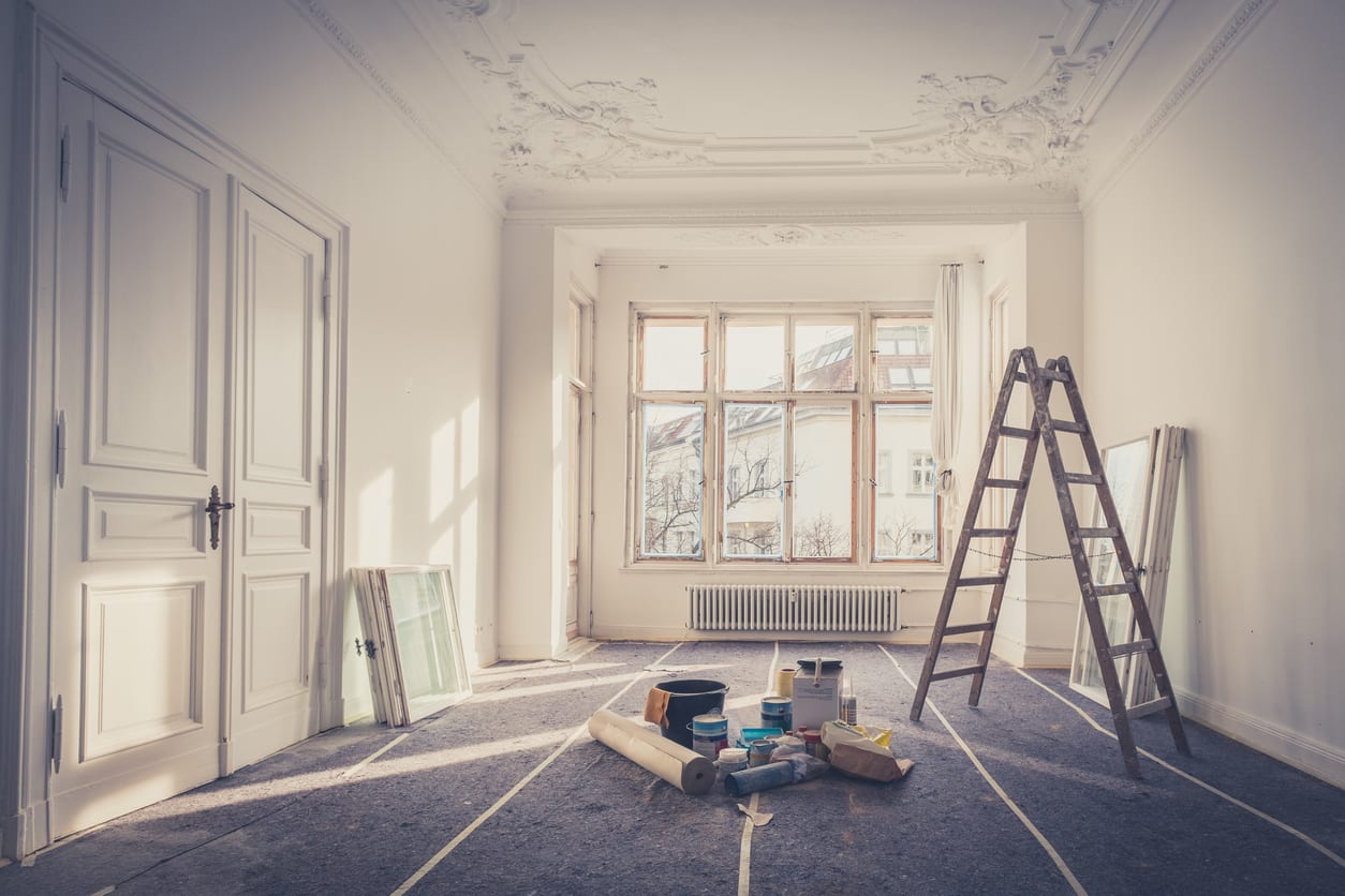 Large, empty room under renovation with ornate ceiling, ladder, and painting supplies on the floor. Light streams through tall windows.