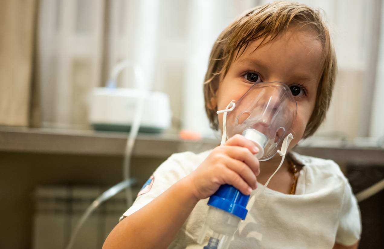 A young child is using a nebulizer mask for breathing treatment indoors, with a nebulizer machine in the background.