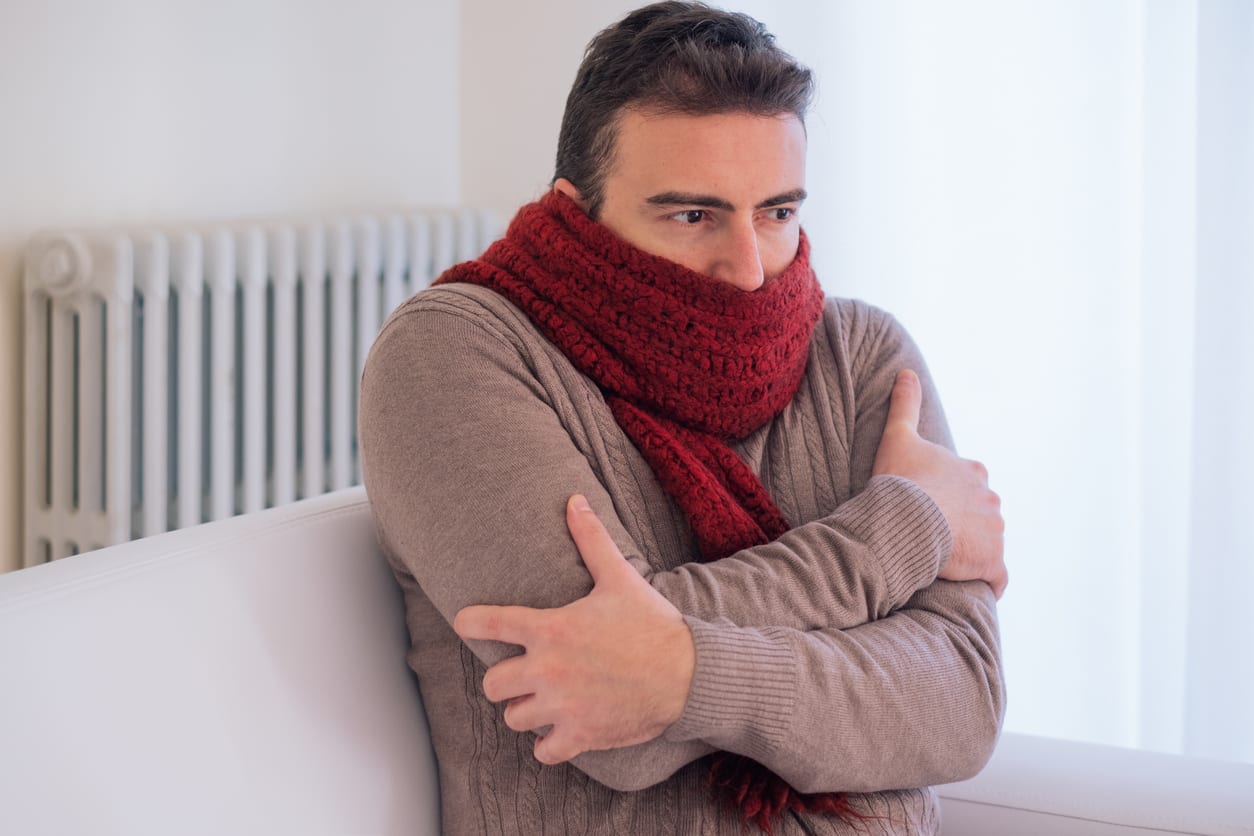 Man sitting on a couch, wrapped in a thick red scarf, with arms crossed and a shivering expression. Radiator in the background.