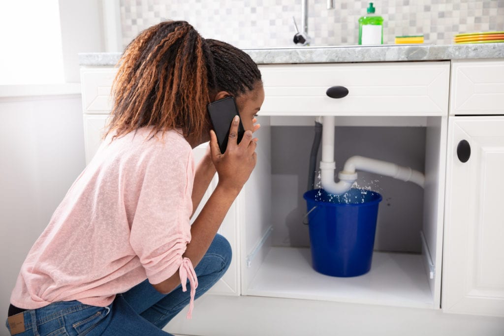 Woman Calling Plumber In Front Of Leaking Sink Pipe
