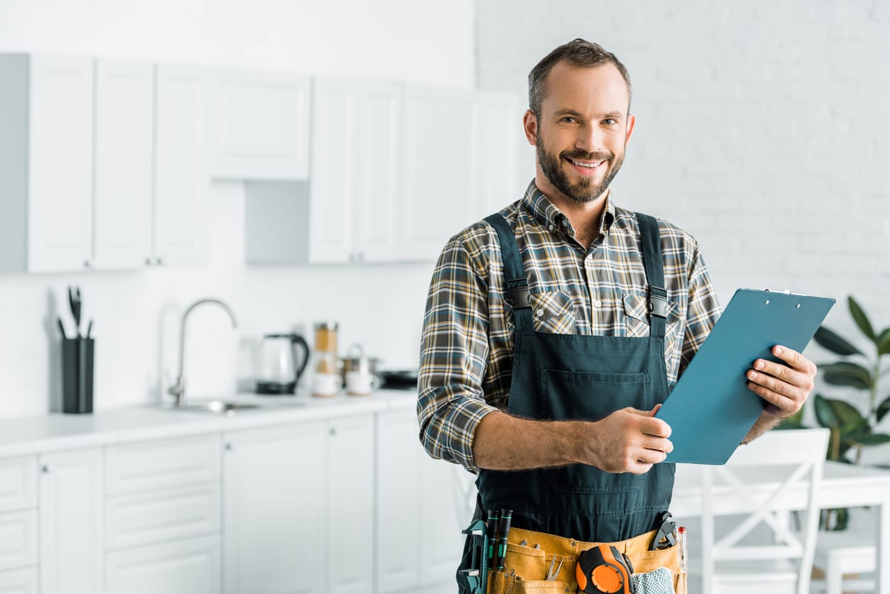 Man in plaid shirt and overalls holds a clipboard while standing in a modern kitchen.