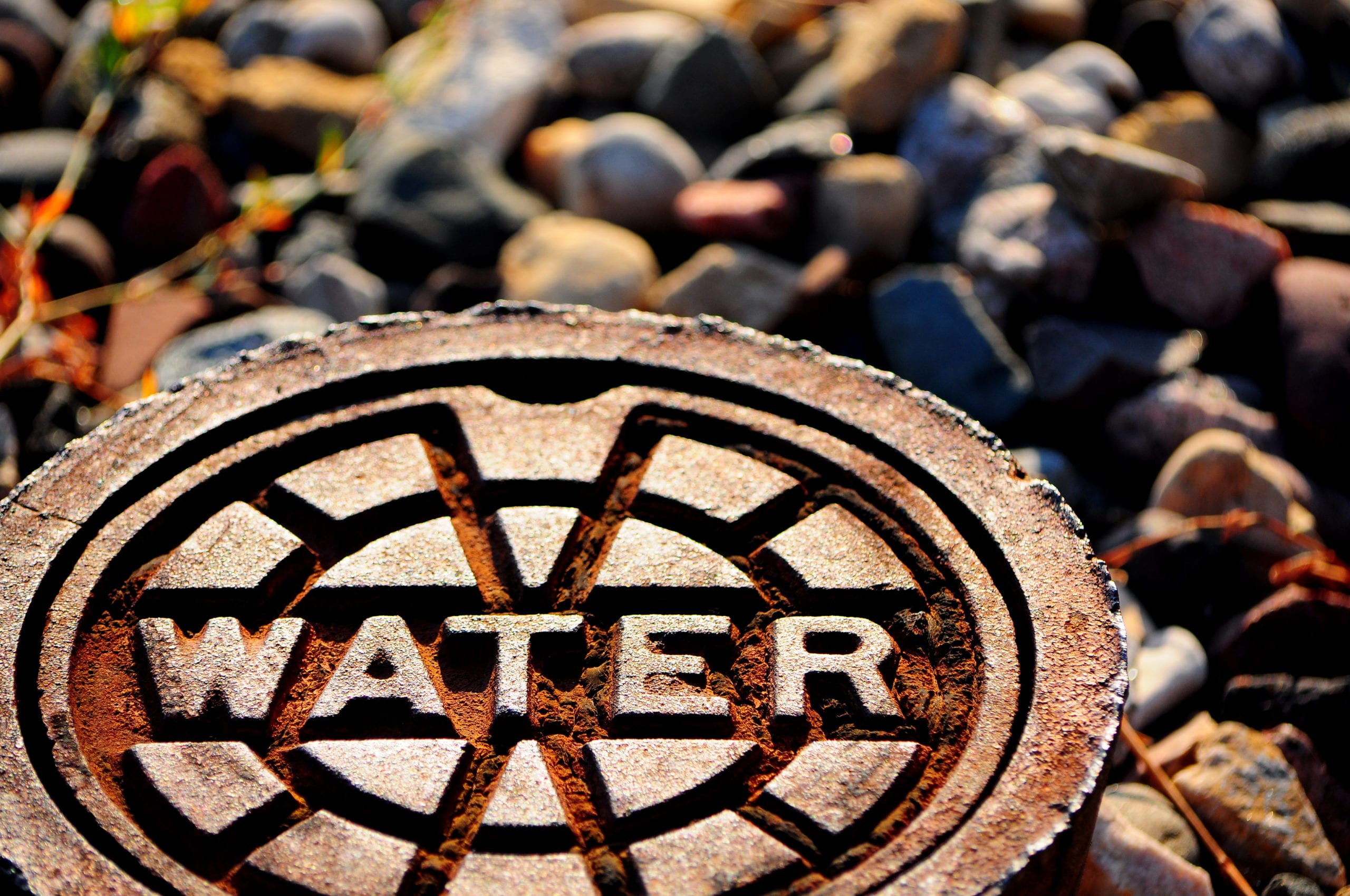 Close-up of a rusted manhole cover labeled "WATER" with a pattern of intersecting lines, surrounded by variously colored smooth stones.