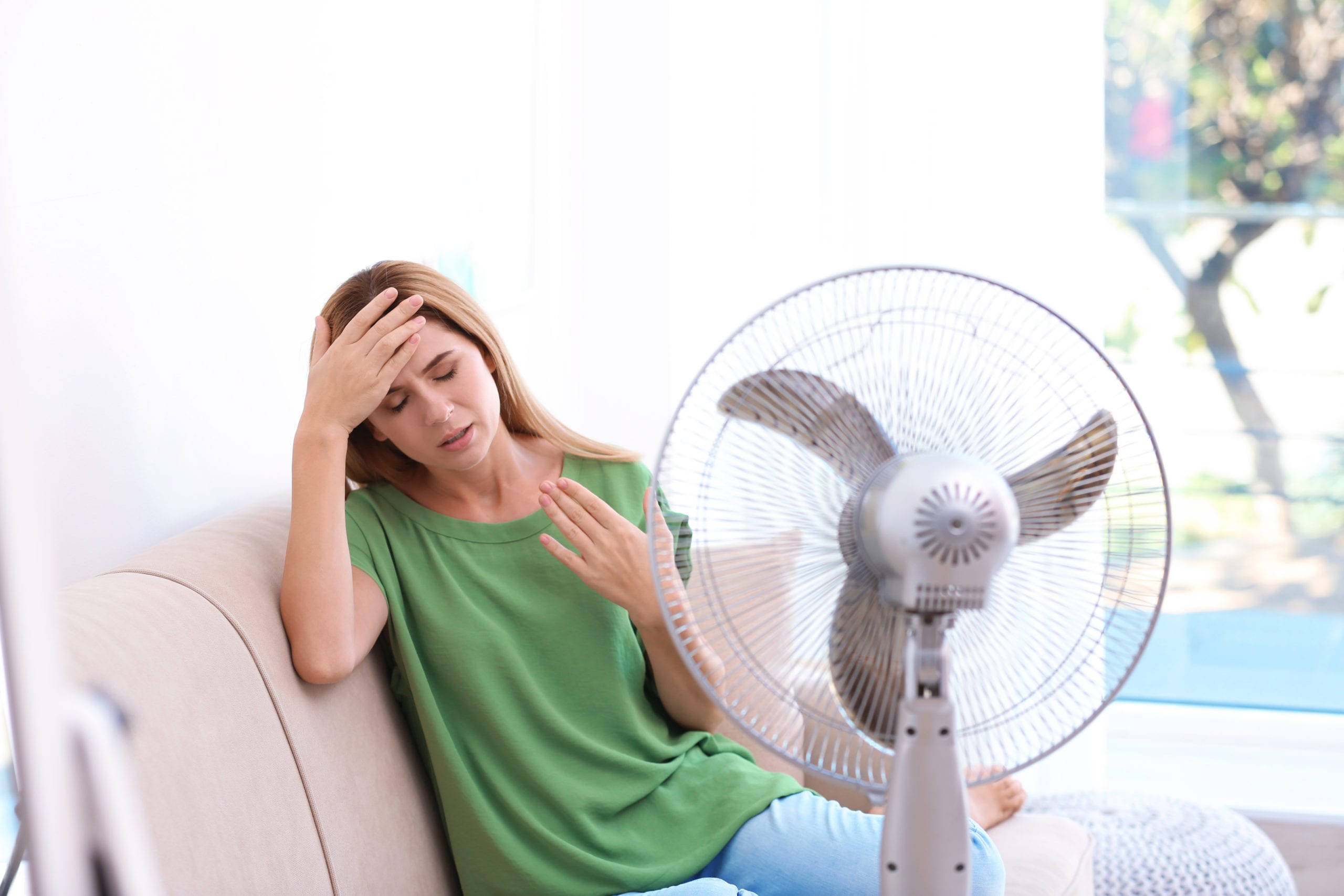 A woman in a green shirt sits on a couch, looking uncomfortable, with one hand on her forehead and the other on her chest. A fan is positioned blowing air towards her.
