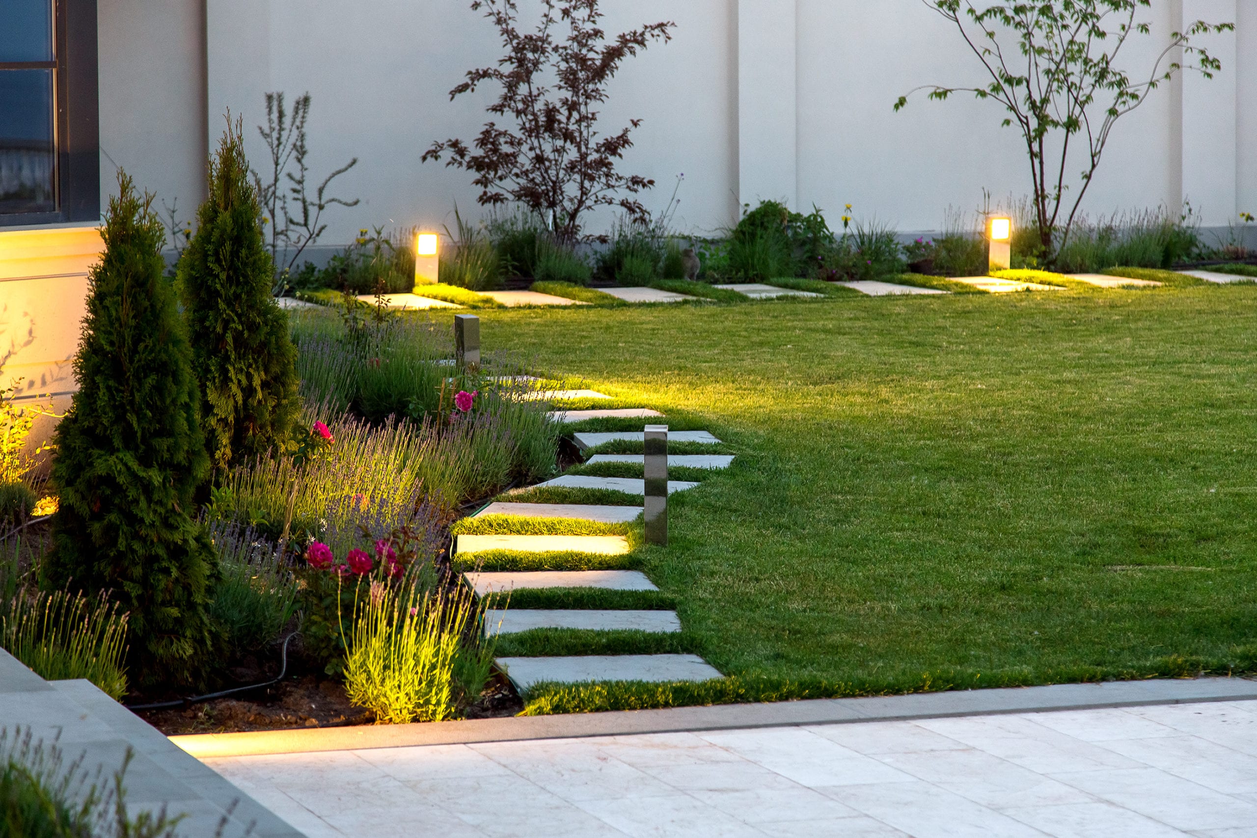 A landscaped garden featuring a stone path with illuminated lights, surrounded by grass and blooming flowers. Trees and bushes are visible near a white building wall.
