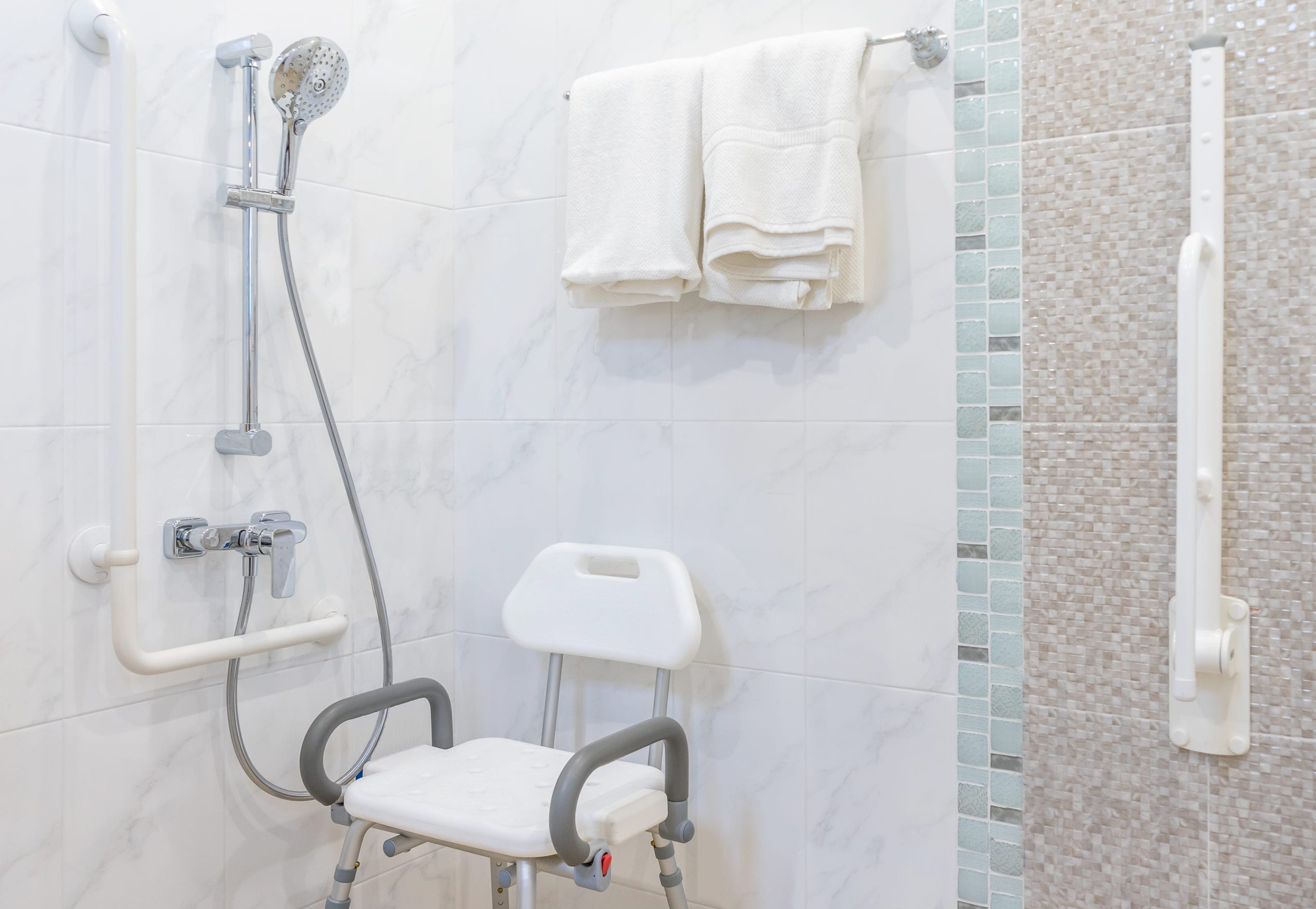 A bathroom with a shower, white tiles, a shower chair with armrests, and folded towels on a rack. A handheld showerhead and safety grab bars are installed on the wall.
