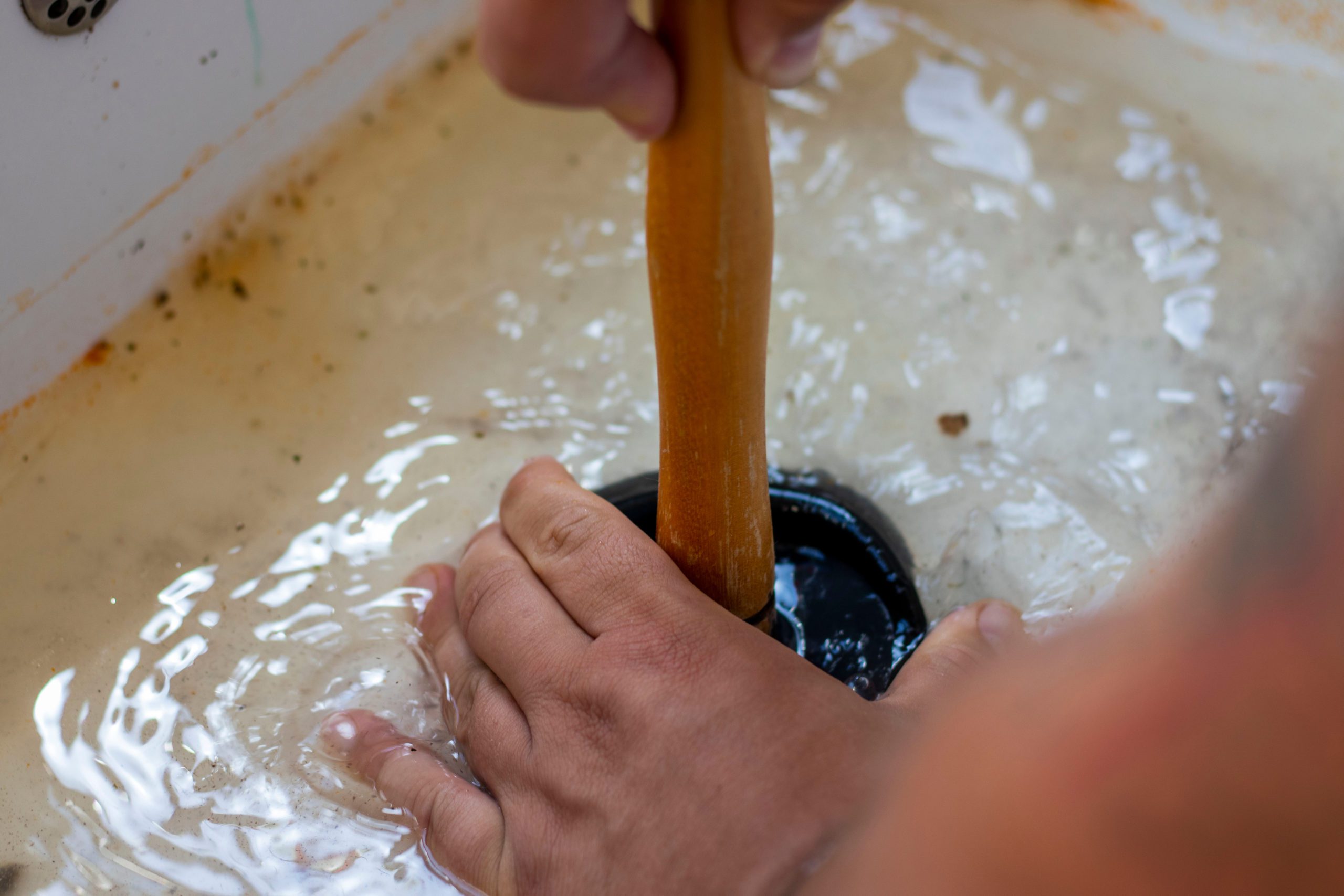 A person using a plunger to clear a clogged sink drain filled with murky water.