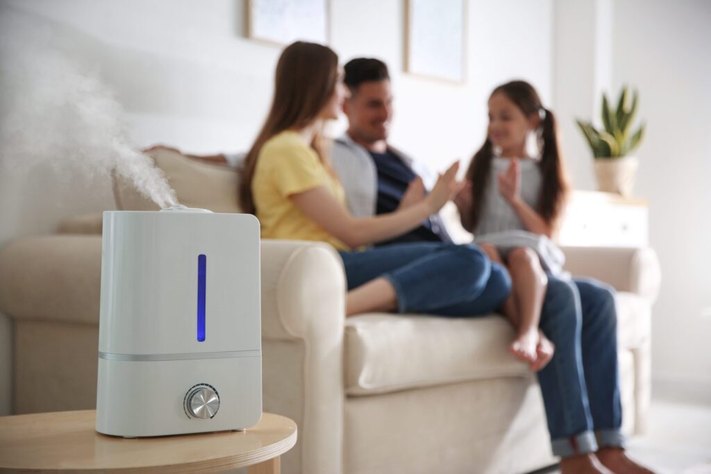 A Humidifier Releases Mist In The Foreground While A Family Of Three Sits On A Sofa In The Background, Chatting And Smiling.