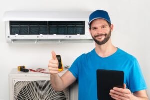 A man in a blue shirt and cap gives a thumbs-up in front of a wall-mounted air conditioning unit, holding a tablet.