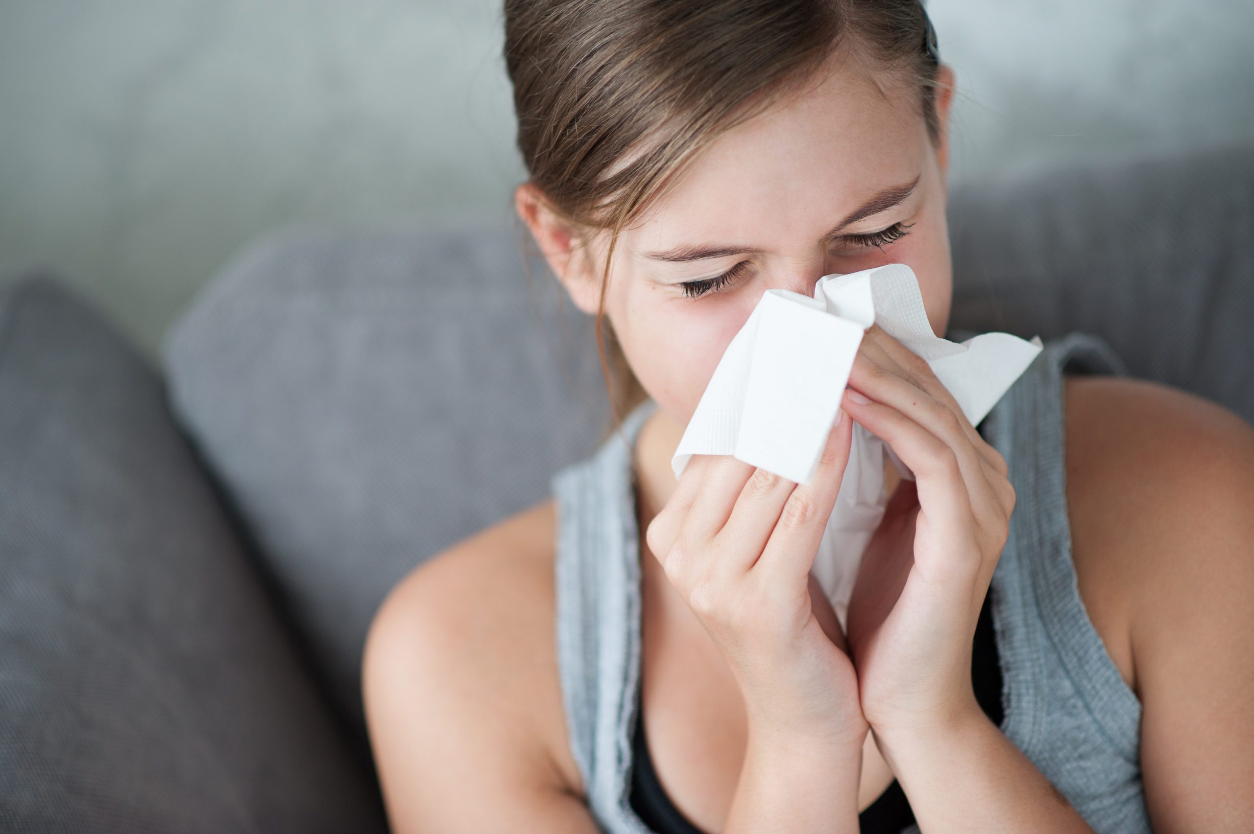 Person sitting on a couch, holding a tissue to their nose, appearing to sneeze.