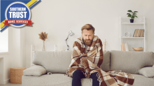 A Man Sitting On A Sofa Wrapped In A Plaid Blanket, Looking Cold. There Is A Logo For Southern Trust Home Services In The Top Left Corner.