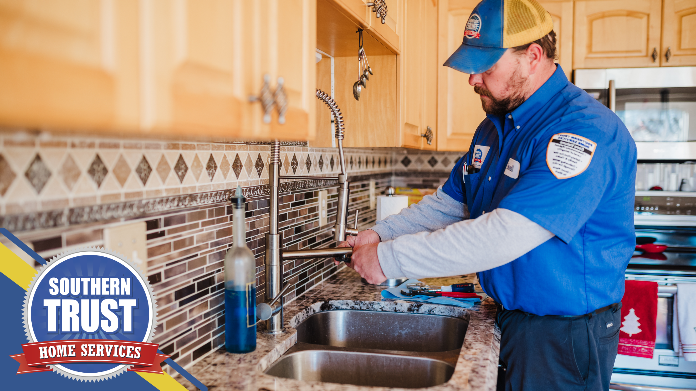 A plumber in a blue uniform is fixing a kitchen faucet in a home. A "Southern Trust Home Services" logo is displayed in the lower left corner.