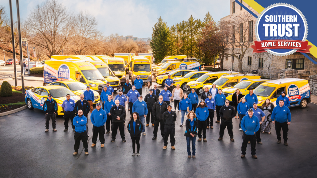 A Group Of People In Blue Shirts Standing In Front Of A Fleet Of Yellow Service Vans With &Quot;Southern Trust Home Services&Quot; Logos In A Parking Lot.