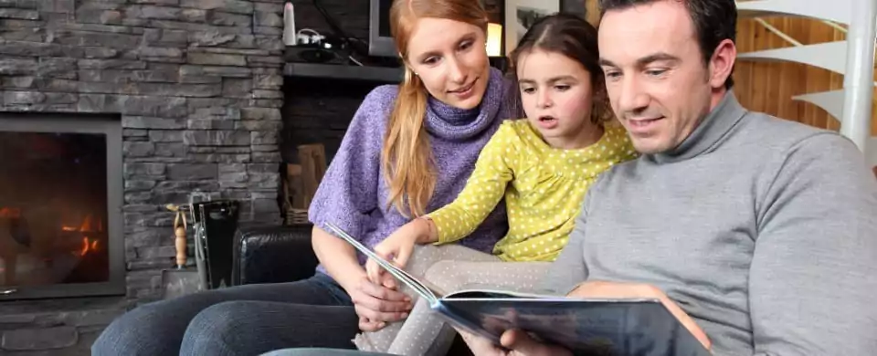 A Man And Woman Sit On A Couch With A Young Girl, Reading A Book Together. A Lit Fireplace Is Visible In The Background, Creating A Cozy Atmosphere That Offers A Perfect Retreat From The Day’s Hustle And Bustle Of Jobs And Careers.