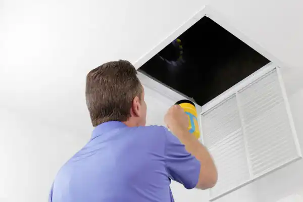 Person in blue shirt shining flashlight into dark ceiling vent, inspecting for potential air duct repair needs.