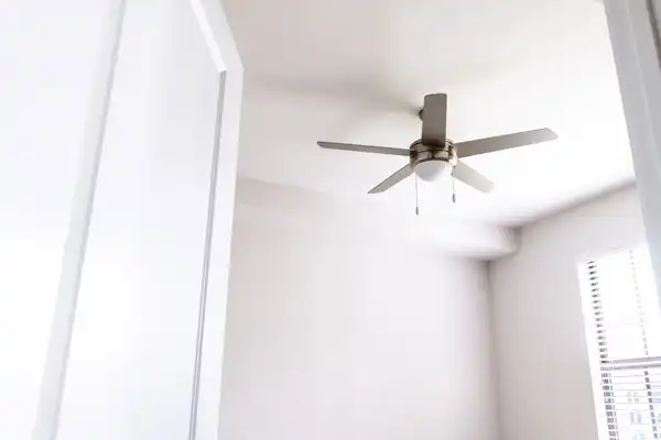 Ceiling fan with five blades and a light fixture gracefully spins in a bright white room, all thanks to expert ceiling fan installation, viewed from a doorway.