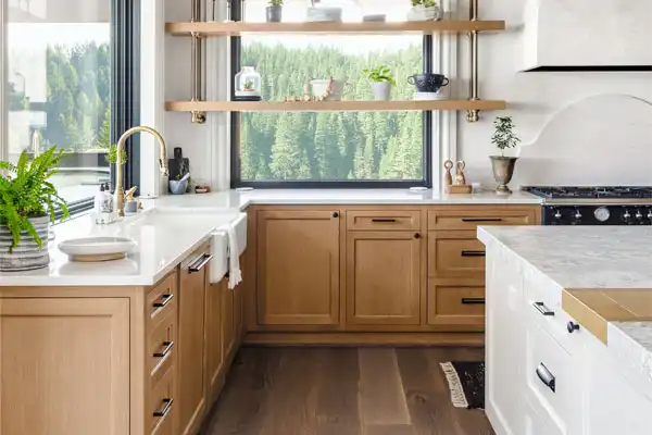 Modern kitchen featuring light wood cabinets, white countertops, and open shelves adorned with plants and kitchenware. A large window offers a view of trees, while the recently completed plumbing repair ensures everything flows seamlessly.
