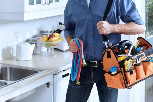 A person with a tool bag and hoses stands in the kitchen, ready for a plumbing repair, near a counter adorned with a plate of fruit and a teapot.