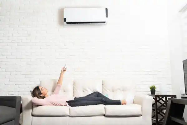 A woman relaxes on a couch, pointing a remote at her newly installed mini-split air conditioner in a bright living room with a stylish white brick wall.