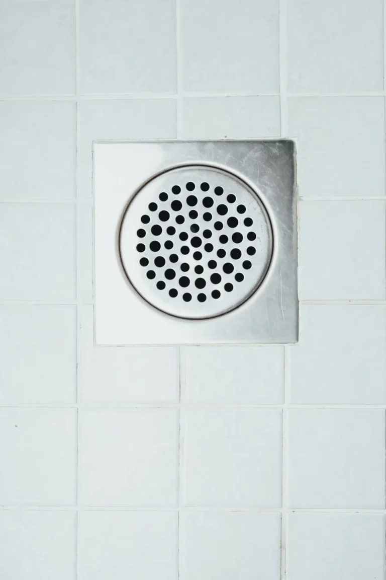 A square metal drain cover with circular holes is seamlessly integrated into the white tiled flooring, ensuring efficient flow and easy access for any necessary rooter service.