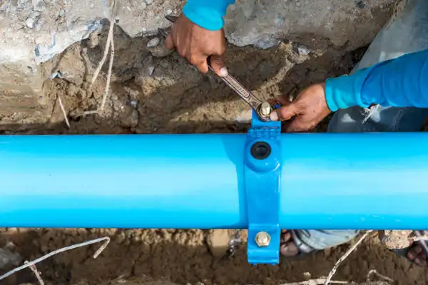 A person focuses intently on Water Line Repair, expertly tightening a bolt on the blue water pipe nestled in the trench with a trusty wrench.