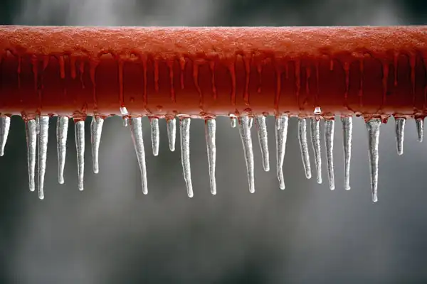 Icicles hanging from a frozen red metal pipe hint at the urgent need for burst pipe repair against a blurred gray background.