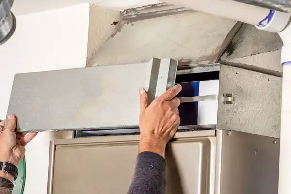 A person skillfully removes a metal panel from the HVAC system, revealing the intricate components within—a crucial step in furnace maintenance.
