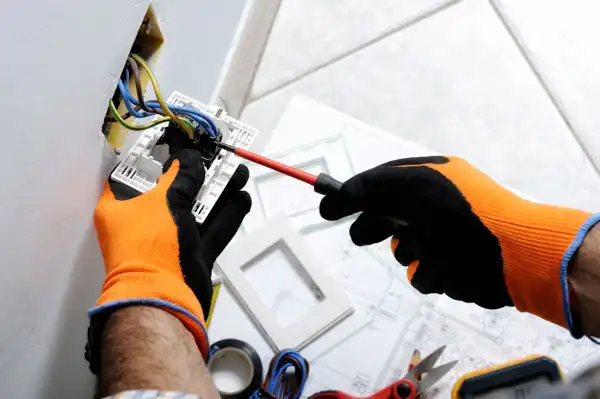 Wearing orange gloves, the person deftly undertakes general electrical repair, fixing wiring in a wall socket with a screwdriver. Nearby, tools and wires are scattered, painting a meticulous scene of precision and skill.