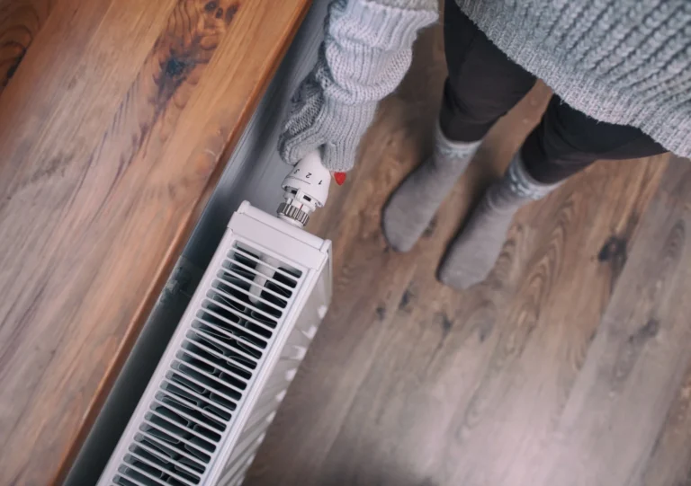Person Wearing Gray Gloves And Socks Adjusts The Thermostat On A Radiator In A Room With Wooden Flooring.