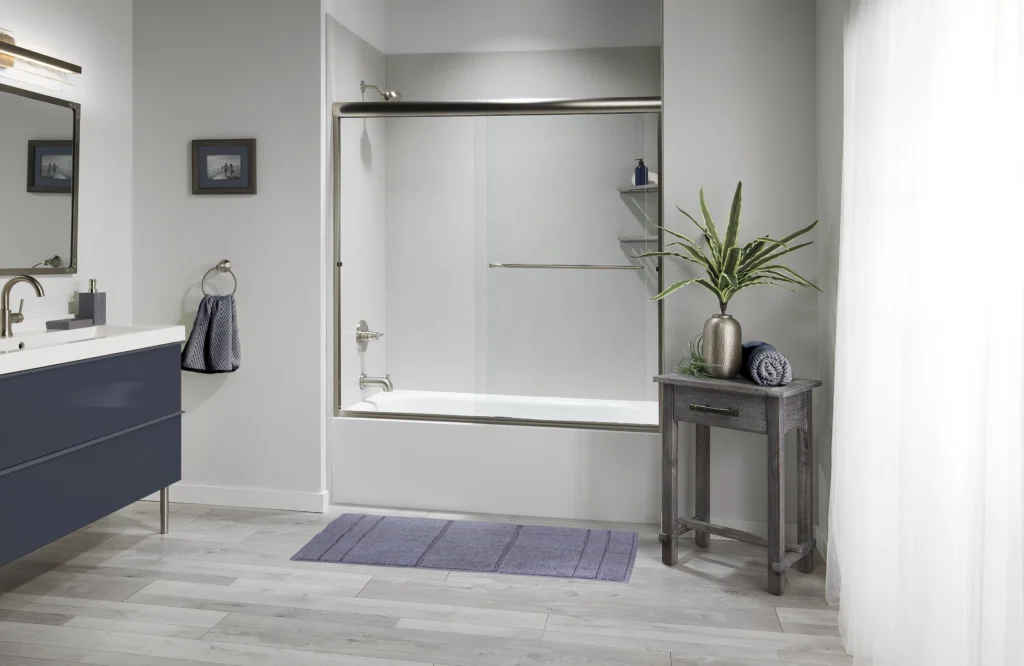 Modern Bathroom With A Glass-Door Shower And Bathtub. A Navy Vanity With A Sink And Mirror Is On The Left. A Small Table With A Plant And Towels Is On The Right. A Purple Bath Mat Lies On The Floor.
