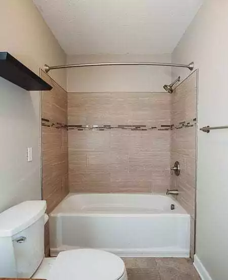 Bathroom With A White Bathtub, Tan Tile Surround, And Silver Shower Fixtures. Toilet On The Left And Towel Rack On The Right Wall.