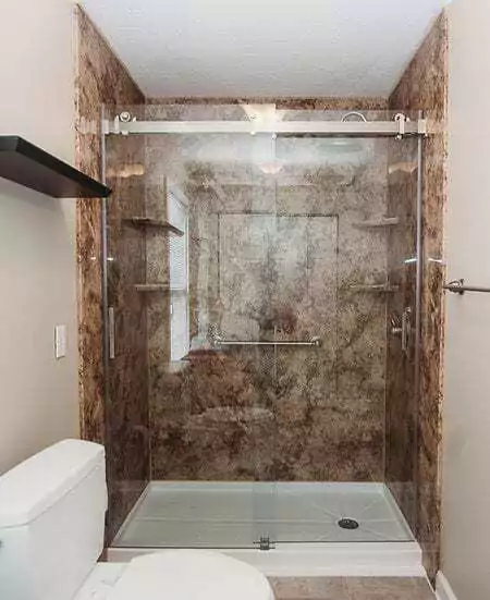 Bathroom With A Brown Marbled Shower Featuring Sliding Glass Doors, Metal Handle, And Wall Shelf. Visible White Toilet With Lid Closed In The Foreground.