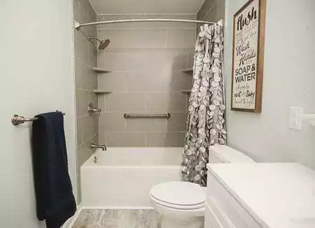 Bathroom With A Shower-Tub Combo, Gray Tile Walls, Patterned Curtain, Towel On A Rack, Framed Sign, And A Toilet Near A White Vanity.