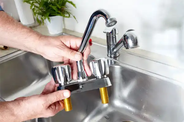 A Person Is Holding A Detached Kitchen Faucet Over A Metal Sink. There Is A Plant In The Background.