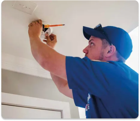 A Person In A Blue Shirt And Cap, Adhering To A Maintenance Plan, Diligently Installs Or Adjusts A Smoke Detector On The Ceiling.